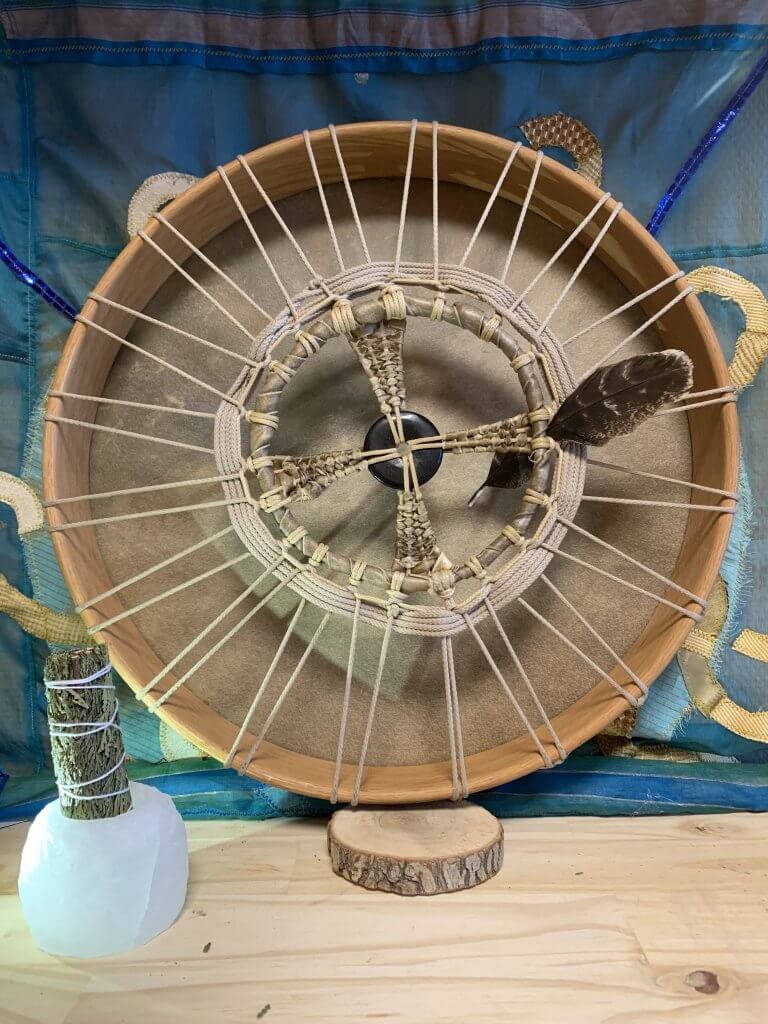 Vue arrière d'un tambour chamanique avec une peau de bison, révélant un système de cordage complexe et méticuleux qui maintient la peau bien tendue sur le cadre circulaire en bois. Les cordes, souvent fabriquées à partir de matières naturelles comme le cuir de bison ou des fibres végétales, traversent le cadre en formant un motif croisé qui allie fonctionnalité et esthétique. Ce réseau de cordage n'est pas seulement crucial pour ajuster la tension de la peau et donc le son produit par le tambour, mais il ajoute également un élément visuel distinctif, avec des nœuds et des torsions qui peuvent servir de décorations ou avoir une signification symbolique dans certaines cultures. L'arrière du tambour peut aussi montrer le soin apporté à la finition du cadre en bois, soulignant l'harmonie entre la force naturelle des matériaux et l'artisanat humain. Cette vue offre une perspective unique sur la construction traditionnelle du tambour, mettant en lumière les aspects techniques et culturels de cet instrument sacré