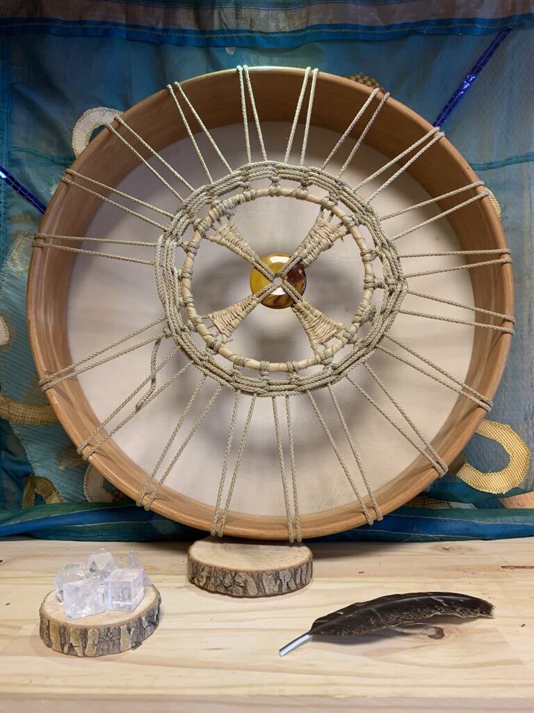 Vue de l'arrière d'un tambour chamanique traditionnel, mettant en évidence le système complexe de cordage qui maintient la peau de chèvre tendue sur le cadre. Les cordes, généralement en matière naturelle comme le cuir ou la fibre végétale, sont méticuleusement entrelacées à travers le cadre en bois, créant un réseau dense et symétrique. Ce cordage n'est pas seulement fonctionnel, assurant la tension nécessaire à la peau pour une qualité sonore optimale, mais présente aussi un aspect esthétique, avec des nœuds et des boucles formant des motifs décoratifs. L'attention portée aux détails dans la conception du cordage reflète le soin et l'intention investis dans la fabrication du tambour, soulignant son importance dans les pratiques chamaniques