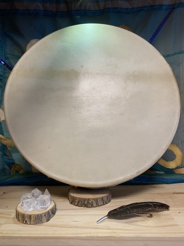 Image d'un tambour chamanique traditionnel circulaire, avec une peau de chèvre tendue sur le cadre. La peau de chèvre, choisie pour sa qualité sonore et sa résistance, est bien fixée autour du cadre en bois, montrant des nuances de couleur naturelle allant du beige clair au brun. Des motifs tribaux ou spirituels peuvent être gravés ou peints sur le cadre, ajoutant une dimension esthétique et culturelle. Le tambour est accompagné d'une mailloche fabriquée avec soin, utilisée pour jouer et invoquer des sons profonds et résonnants lors de cérémonies chamaniques.