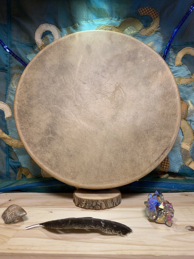 Vue de face d'un tambour chamanique traditionnel orné d'une peau de veau tendue avec soin sur un cadre circulaire. La peau de veau, sélectionnée pour sa texture lisse et sa capacité à produire des sons clairs et profonds, présente une teinte naturelle allant du crème au brun clair, avec des variations subtiles qui témoignent de l'authenticité du matériau. Au centre du tambour, le point focal est souvent souligné par un marquage ou reste naturel, servant de guide pour le joueur et symbolisant le cœur ou l'esprit du tambour dans de nombreuses traditions. Autour du bord, la peau est attachée de manière experte au cadre en bois, garantissant une tension parfaite pour une résonance optimale. Cette face avant invite à la contemplation et à la connexion spirituelle, chaque coup de mailloche évoquant des sons enveloppants qui peuvent faciliter la méditation ou le voyage chamanique